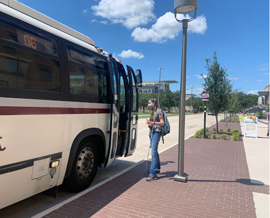A young white woman with a white cane boarding a public bus. She is wearing a mask and a backpack.